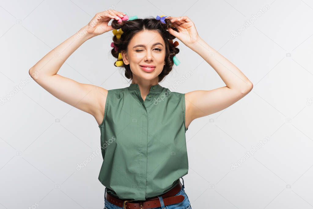 cheerful young woman with colorful hair curlers winking eye isolated on grey