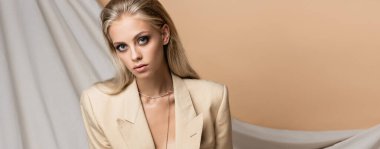 blonde woman in blazer looking at camera on beige draped background, banner clipart