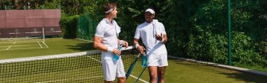 Gülümseyen adam Afrika kökenli Amerikalı bir arkadaşın yanında tenis kortunda, pankartta 