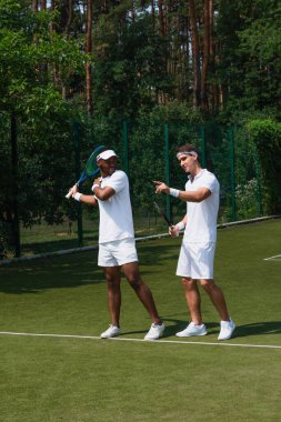 Parmakla işaret eden sporcu, kortta tenis raketi olan Afrikalı Amerikalı bir arkadaşın yanında. 
