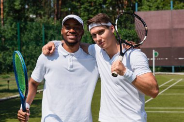 Tenis raketleriyle tenis kortunda sarılan çok ırklı sporcular. 