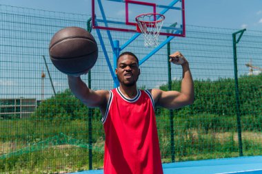 Afrikalı Amerikalı sporcu kaslarını gösteriyor ve elinde basketbol topu tutuyor. 