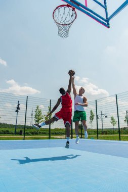 Wide angle view of interracial men jumping while playing streetball outdoors  clipart