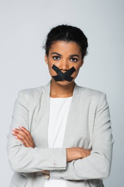 Ağzı bantlı, kolları çaprazlanmış bir şekilde gri üzerinde duran Afro-Amerikan iş kadını.