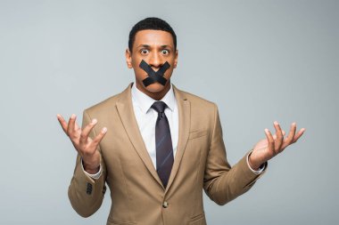 Kafası karışmış Afrikalı Amerikalı iş adamı. Ağzında bant var. Gri üzerinde izole edilmiş el kol hareketi yapıyor.