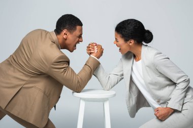 side view of african american businesswoman and man doing arm wrestling on white chair isolated on grey clipart
