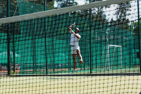 Smiling african american player with tennis racket jumping near blurred net 