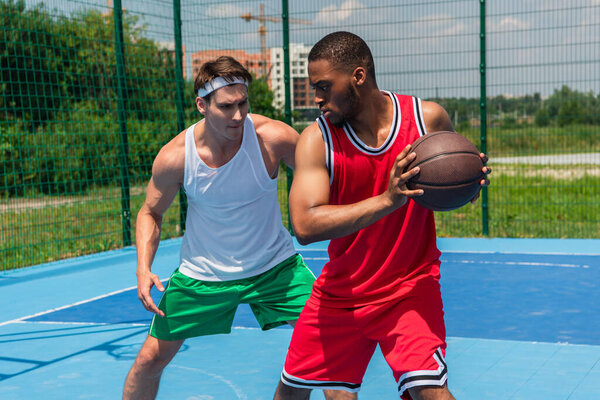 African american streetball player holding ball near friend on playground 