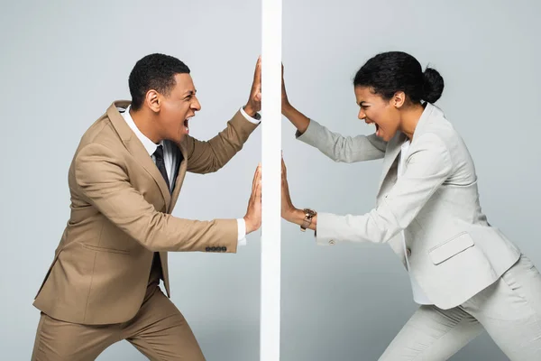 irritated african american businessman and businesswoman pulling wall and screaming on grey, gender equality concept