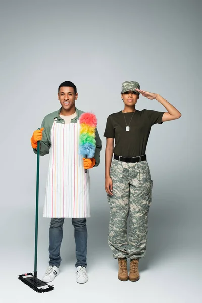 full length of female soldier in cap near male cleaner in apron with mop and dust brush on grey