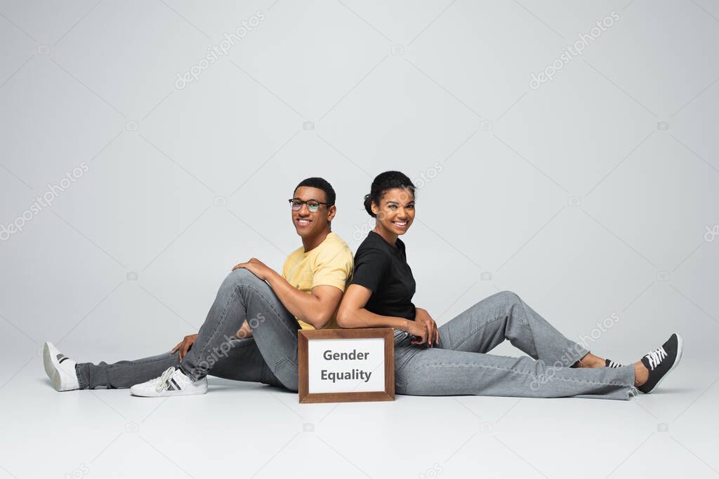 full length of happy african american man and woman sitting near placard with gender equality lettering on grey