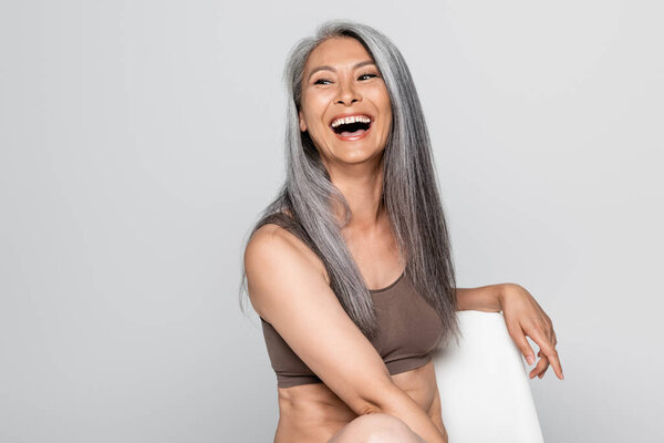 Excited asian woman in bra sitting on chair isolated on grey