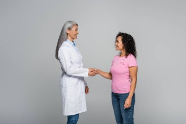 Side view of asian doctor and african american patient with pink ribbon shaking hands isolated on grey clipart