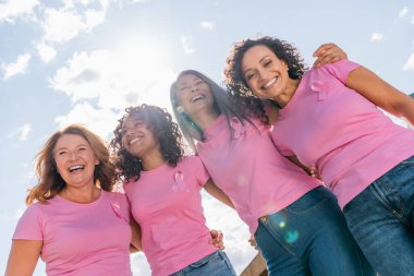 Low angle view of cheerful multicultural women with pink ribbons hugging outdoors  clipart