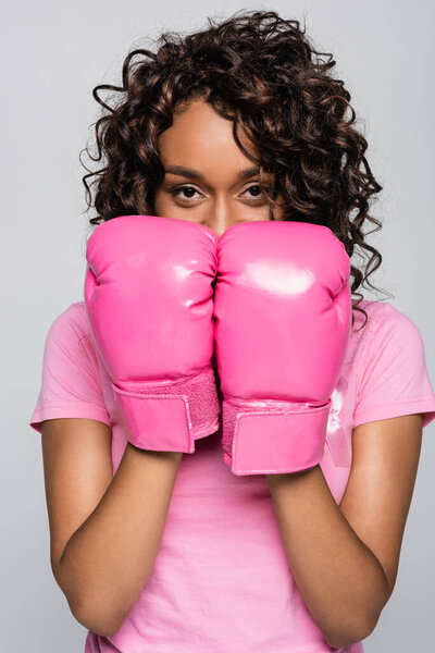 African american woman with pink ribbon and boxing gloves looking at camera isolated on grey