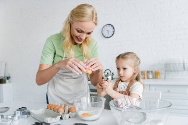Neşeli kadın mutfakta kızıyla yemek pişirirken yumurtayı kaseye kırıyor.