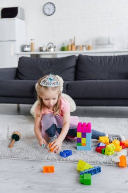 kid in toy crown playing with multicolored building blocks on floor at home clipart