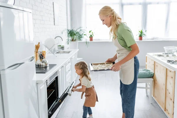 Vista Lateral Menina Abrindo Forno Elétrico Perto Mãe Com Biscoitos — Fotografia de Stock