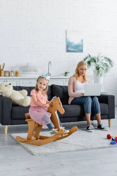 Child Toy Crown Riding Rocking Horse Blurred Mom Working Laptop — Stock Photo, Image
