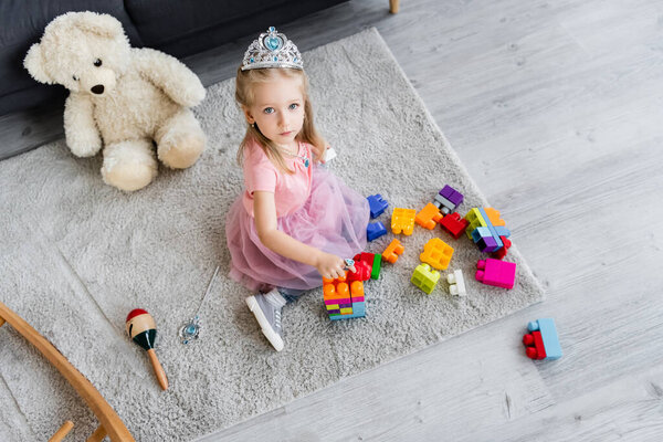 child in princess costume sitting on floor carpet near toys and teddy bear