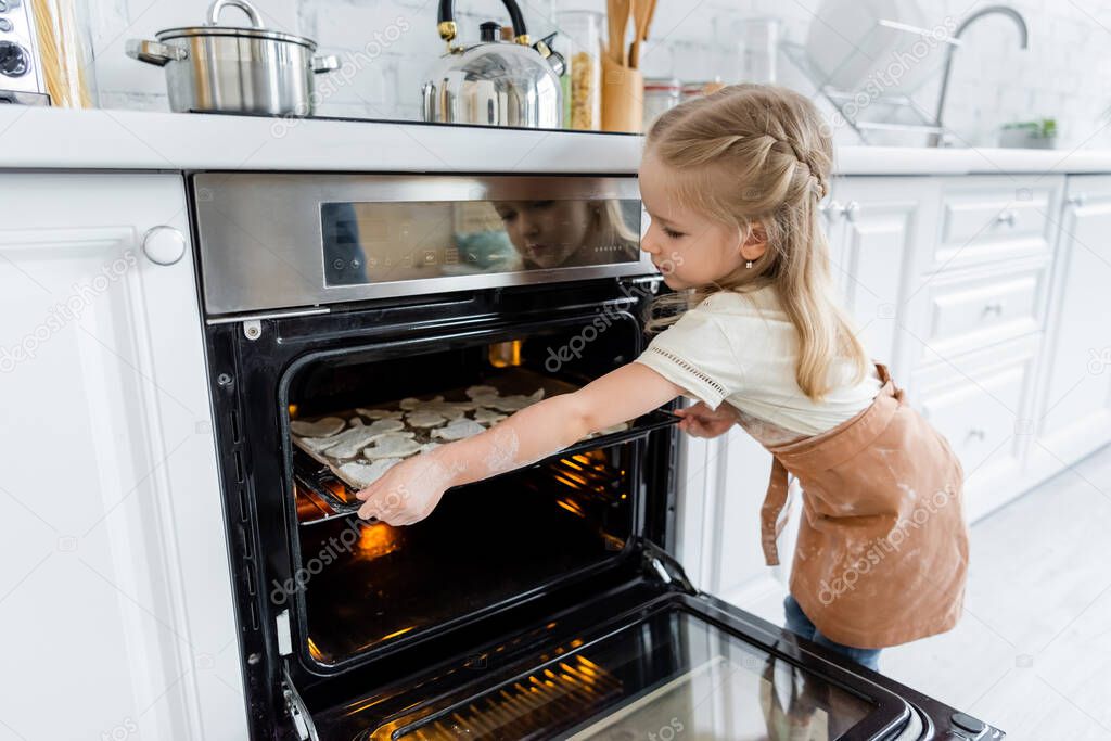 girl in apron placing baking sheet with cookies in oven