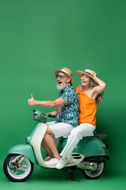 Orta yaşlı mutlu bir adam, yeşil motosikletli Asyalı eşiyle gezerken baş parmağını gösteriyor. 
