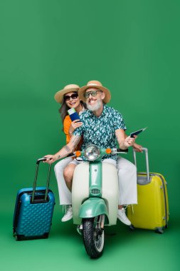Neşeli orta yaşlı ve çok ırklı güneş şapkalı çift ellerinde pasaport ve bavullarıyla yeşil motosiklet sürerken