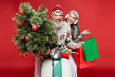 Neşeli orta yaşlı bir adam Asyalı bir eşin yanında Noel ağacını tutarken kırmızı motosiklete biniyor. 