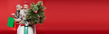 Heyecanlı orta yaşlı bir adam Asyalı karısının yanında Noel ağacı tutarken kırmızı pankartta motosiklet sürüyor.