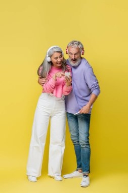 Uzun, mutlu, orta yaşlı, kablosuz kulaklıklı bir kadın akıllı telefonuyla şaşırmış kocanın yanındaki sarıyı işaret ediyor.