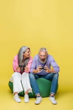 KYIV, UKRAINE - 10 AĞUSTOS 2021: Irklar arası ve orta yaşlı kadın sırıtan dişleri ile sarı üzerine şaşırmış kocasıyla video oyunu oynarken 