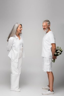full length of happy middle aged man hiding bouquet of flowers behind back near asian wife on grey clipart
