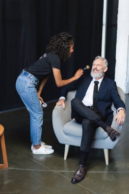 Esmer Afrikalı Amerikalı makyöz röportaj stüdyosunda mutlu iş adamına yüz pudrası uyguluyor.