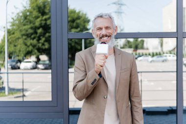 Blazer ceketli neşeli muhabir mikrofon tutuyor ve binanın yakınında haber yapıyor. 