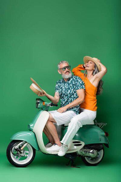 happy and multiethnic middle aged woman holding sun hat while riding moped with husband on green 