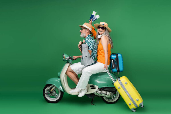 cheerful middle aged asian woman holding passports near husband holding flippers while riding motor scooter on green