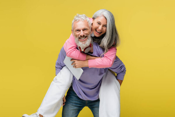 cheerful middle aged man piggybacking happy asian wife with digital tablet isolated on yellow 