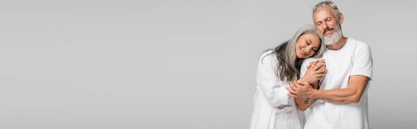 счастливая межрасовая и зрелая пара с закрытыми глазами, держась за руки и обнимаясь изолированно на сером, баннер