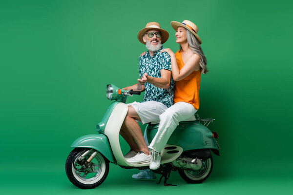 cheerful asian woman in sun hat and middle aged husband riding moped on green 