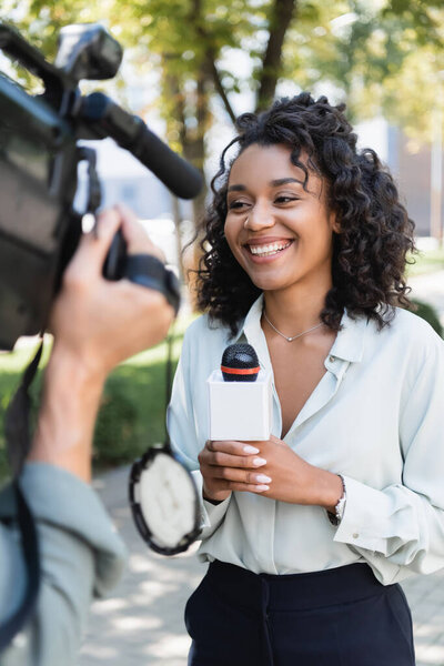 smiling african american journalist with microphone doing reportage near blurred cameraman with video camera 