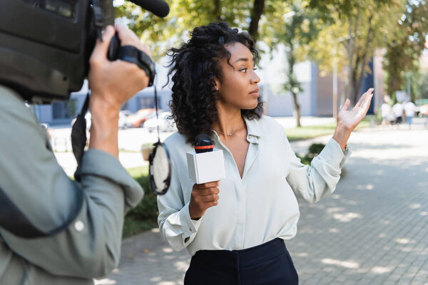 pretty african american journalist with microphone pointing with hand while doing reportage near blurred cameraman