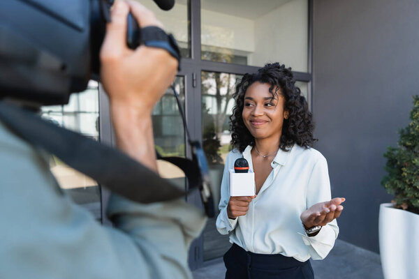 happy african american journalist with microphone gesturing while doing reportage near blurred cameraman