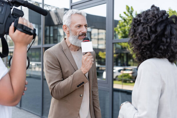 reporter with microphone interviewing african american woman near blurred cameraman with video camera