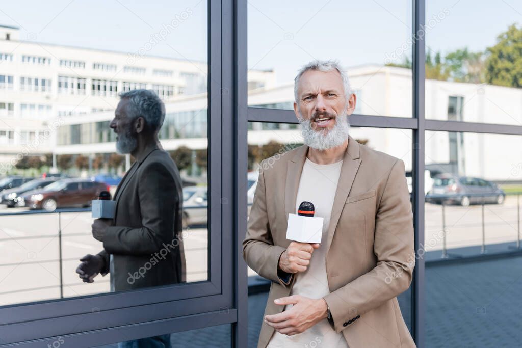 bearded reporter in blazer holding microphone and making reportage near building 