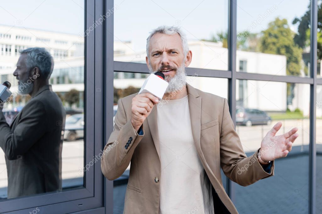 bearded reporter in blazer holding microphone and talking near building 