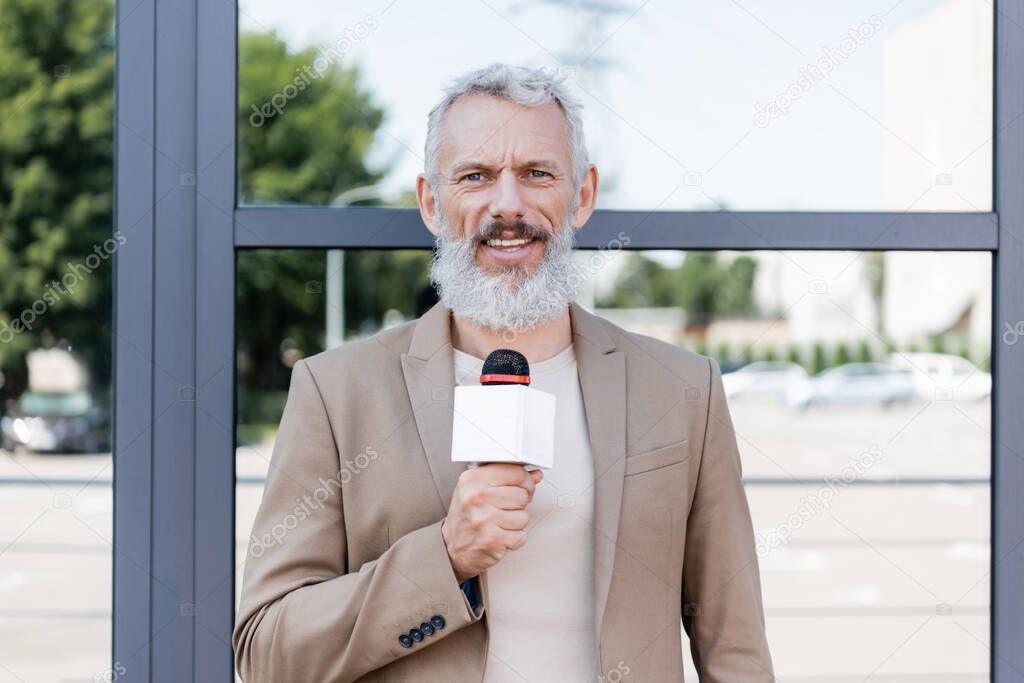 bearded announcer in blazer holding microphone and making reportage near building 