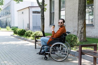 Genç engelli adam tekerlekli sandalyede dizüstü bilgisayarıyla oturuyor ve dışarıda içki ısmarlıyor.