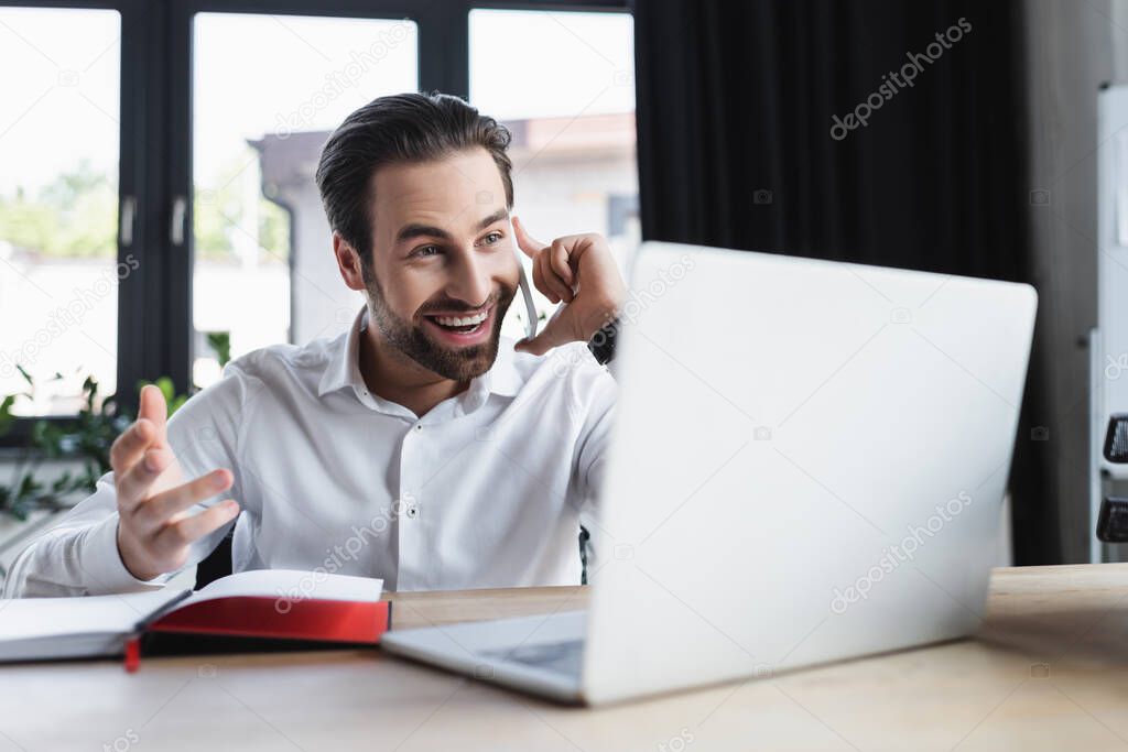 happy businessman gesturing while talking on smartphone near blurred laptop in office