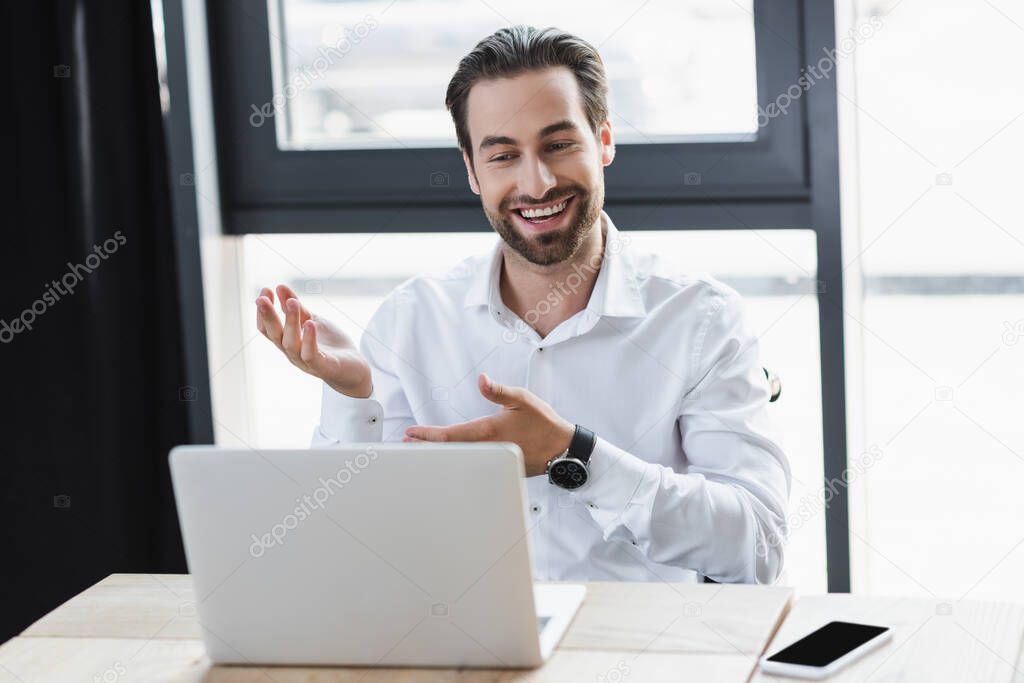 positive businessman pointing with hands near laptop and smartphone with blank screen in office