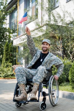 Küçük Amerikan bayrağı taşıyan neşeli asker el kaldırmış dışarıdaki kameraya bakıyor.